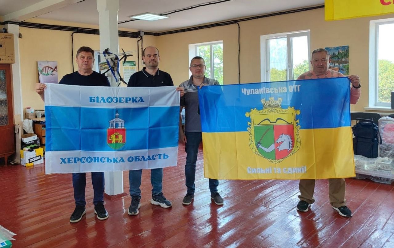 Четверо чоловіків стоять у кімнаті з дерев'яною підлогою та світло-жовтими стінами, тримаючи два великі прапори. Чоловіки ліворуч тримають біло-блакитний прапор із написом 