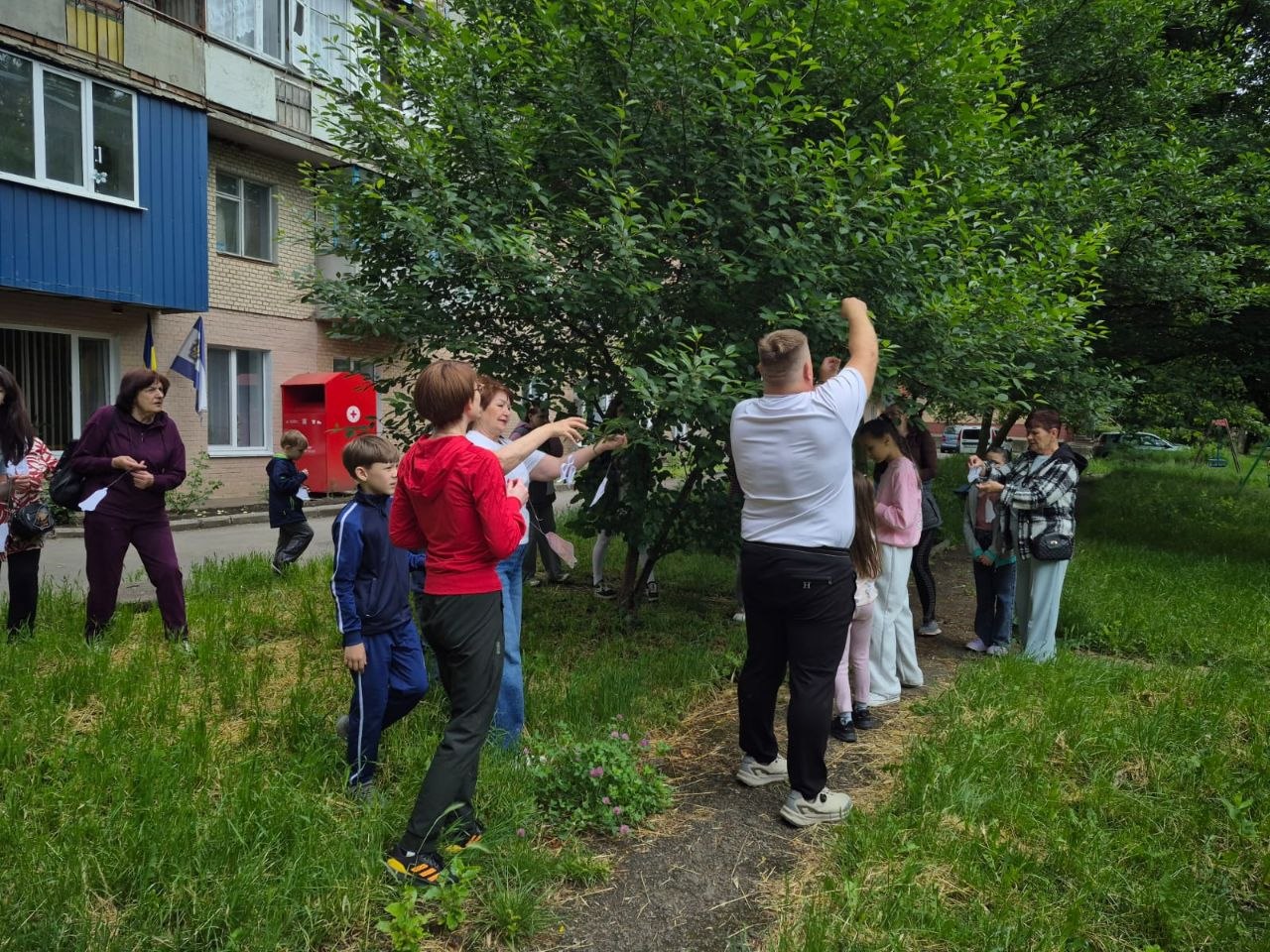 Загальний план подвір'я біля багатоповерхового будинку, де проходить меморіальна акція. Дорослі та діти різного віку розійшлися навколо великого дерева, прикрашаючи його гілки паперовими виробами. На задньому плані видно частину будівлі зі світлою цеглою та синім балконом, а також червоний металевий бокс із медичним логотипом (символом Червоного Хреста).