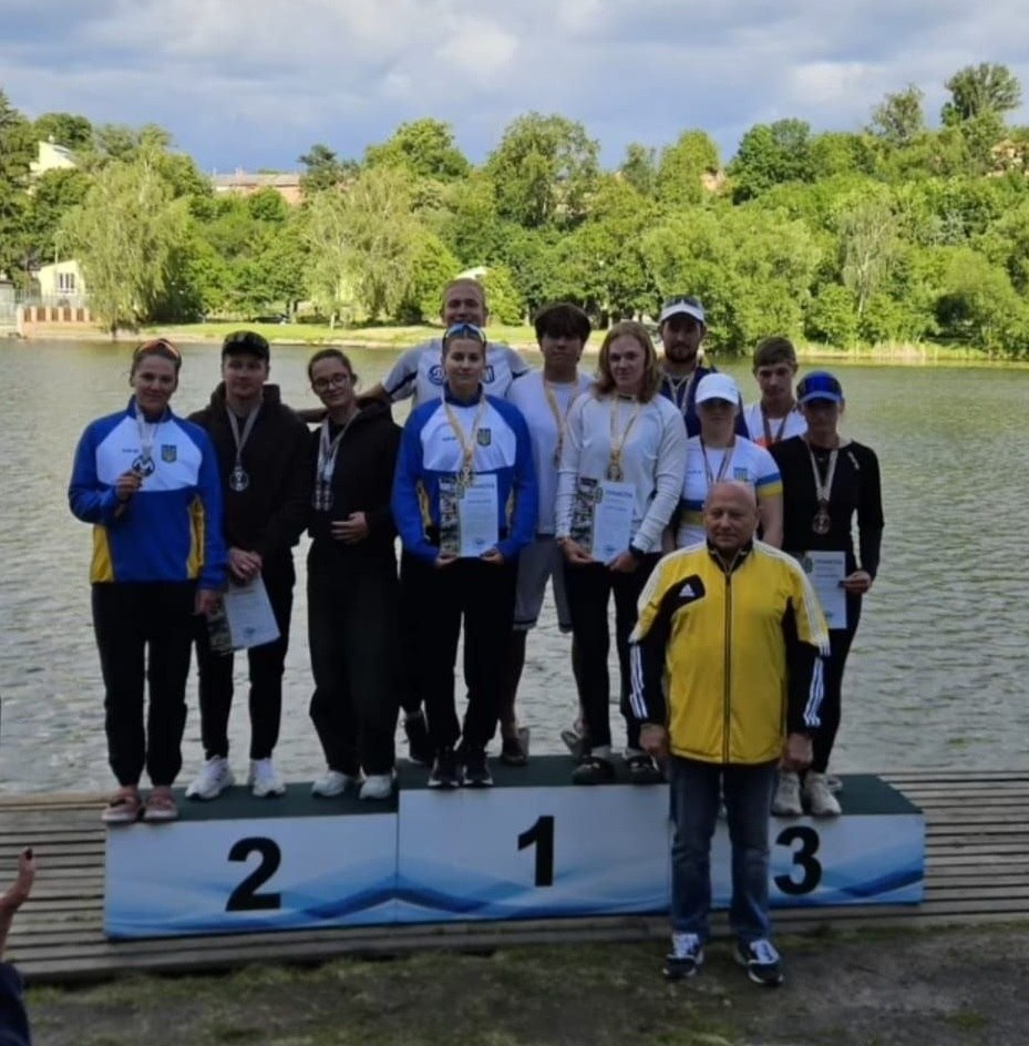 Група спортсменів (чоловіки та дівчата) з медалями та грамотами стоїть на трирівневому спортивному п'єдесталі на дерев'яному помості біля річки або озера. Переможці на першій сходинці тримають у руках нагороди, дехто одягнений у синьо-жовту спортивну форму з гербом України. На передньому плані праворуч стоїть літній чоловік у жовто-чорній спортивній куртці та синіх джинсах. На задньому плані — протилежний зелений берег із деревами та поодинокими будівлями під хмарним небом.