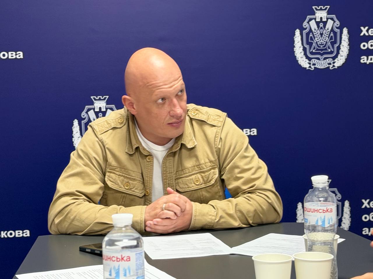 Альтернативний текст: Чоловік військового чиновницького вигляду із поголеною головою, вдягнений у тактичну сорочку кольору хакі поверх білої футболки, сидить за столом на тлі синього банера ХОВА. Його руки складені в замок, погляд спрямований убік під час обговорення. На столі перед ним лежать папери.