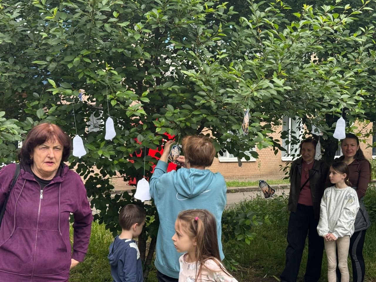 Жінка у блакитному худі з капюшоном закріплює паперову ілюстрацію у формі церковного дзвона на гілці зеленого дерева. Навколо неї на нитках уже розвішані інші білі паперові дзвіночки. Ліворуч на передньому плані стоїть жінка у темно-бордовій кофті, яка дивиться вбік, а праворуч на задньому плані — літня жінка, дівчинка та інша учасниця акції.