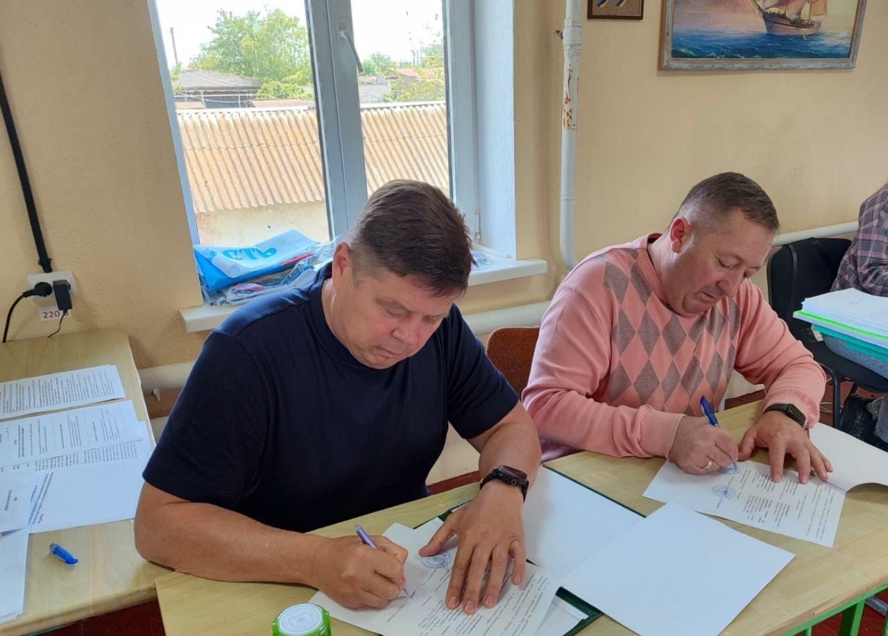 Двоє чоловіків сидять за дерев'яним столом біля вікна в офісному приміщенні та підписують документи синіми ручками. Чоловік ліворуч у темно-синій футболці, чоловік праворуч у рожевому светрі з ромбоподібним візерунком. На столі лежать кілька аркушів паперу, на деяких з яких видно текст, а один аркуш згорнуто у рулон. За вікном видно дах іншої будівлі та зелені дерева. Кімната має світло-бежеві стіни та дерев'яну підлогу.