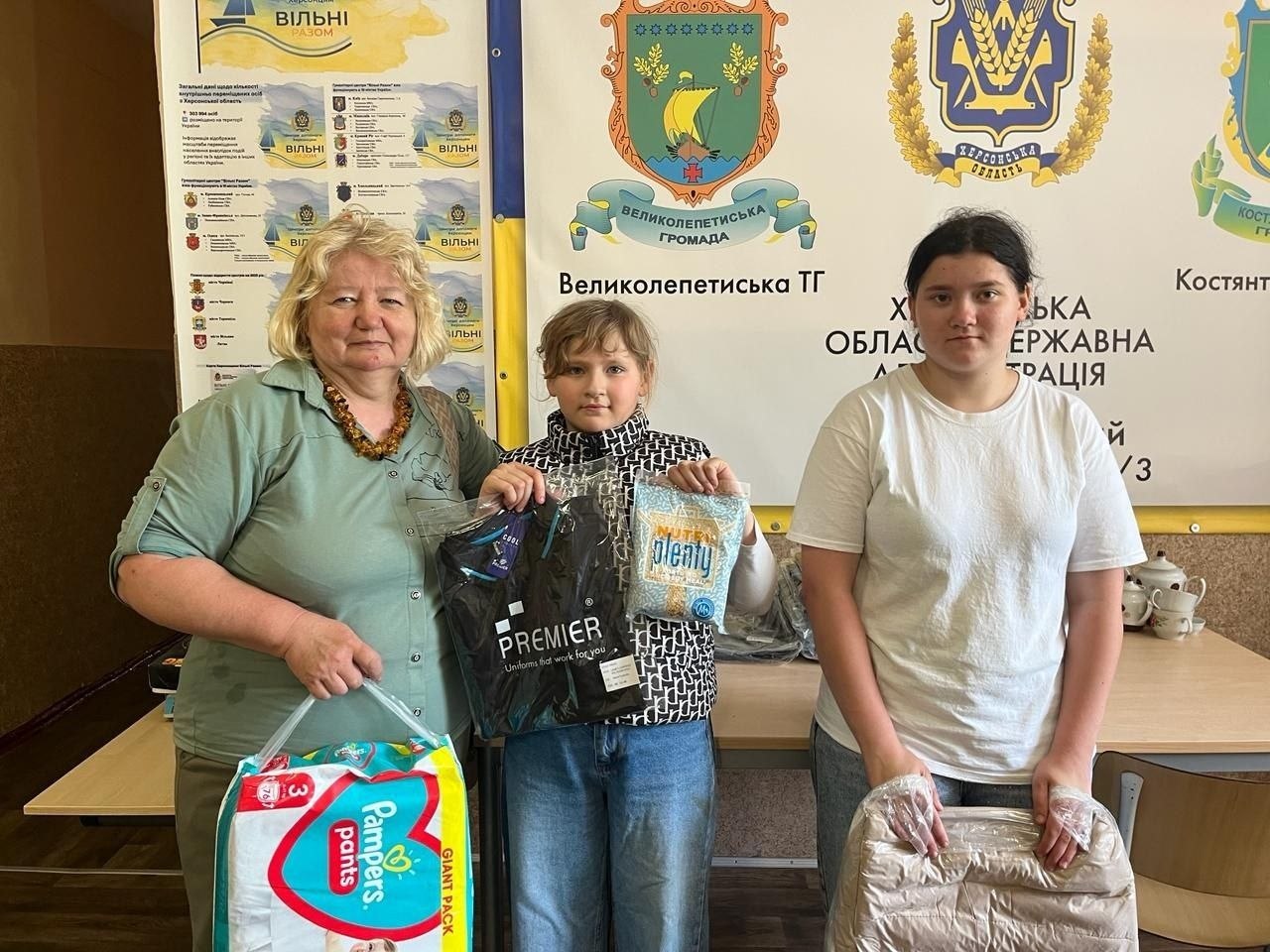 Видача гуманітарної допомоги в офіційному центрі підтримки. Три жінки стоять поруч і тримають отримані речі: велику пачку підгузків Pampers pants, упакований новий одяг та продуктові набори. На задньому плані розміщено інформаційні стенди з гербами Великолепетиської територіальної громади та Херсонської області.