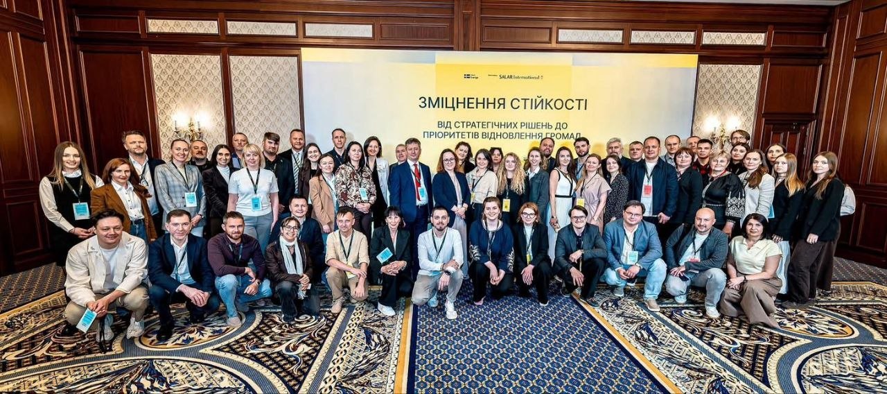 Панорамне групове фото великої кількості учасників офіційного заходу у розкішній залі зі стінами з темного дерева та візерунчастим синьо-бежевим килимом. Кілька десятків людей (чоловіки та дівчата) стоять у кілька рядів, а частина сидить навпочіпки на передньому плані. У багатьох на шиї висять бейджі учасників. По центру позаду них встановлено великий світло-жовтий банер із логотипом «SALAR International» та великим чорним написом українською мовою: «ЗМІЦНЕННЯ СТІЙКОСТІ. ВІД СТРАТЕГІЧНИХ РІШЕНЬ ДО ПРІОРИТЕТІВ ВІДНОВЛЕННЯ ГРОМАД».