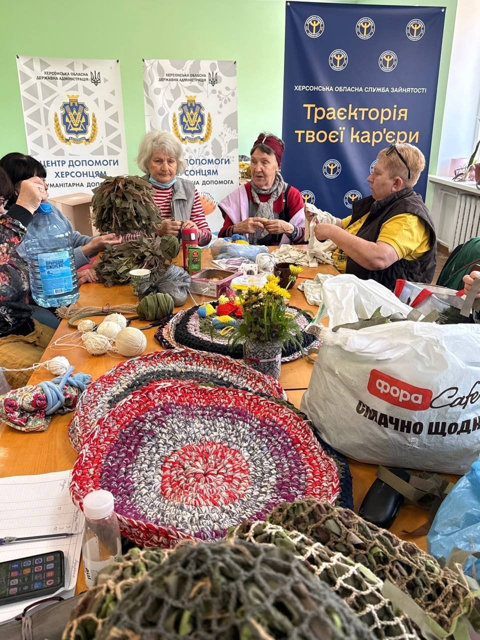 Процес рукоділля волонтерок великим планом. На дерев'яному столі лежать кілька готових круглих килимків ручної роботи, зв'язаних із різнокольорових тканинних смужок, клубки ниток, а також заготовки для маскувальних засобів захисного кольору. На задньому плані три жінки похилого віку спілкуються та працюють, сидячи перед інформаційними стендами хабу допомоги херсонцям.