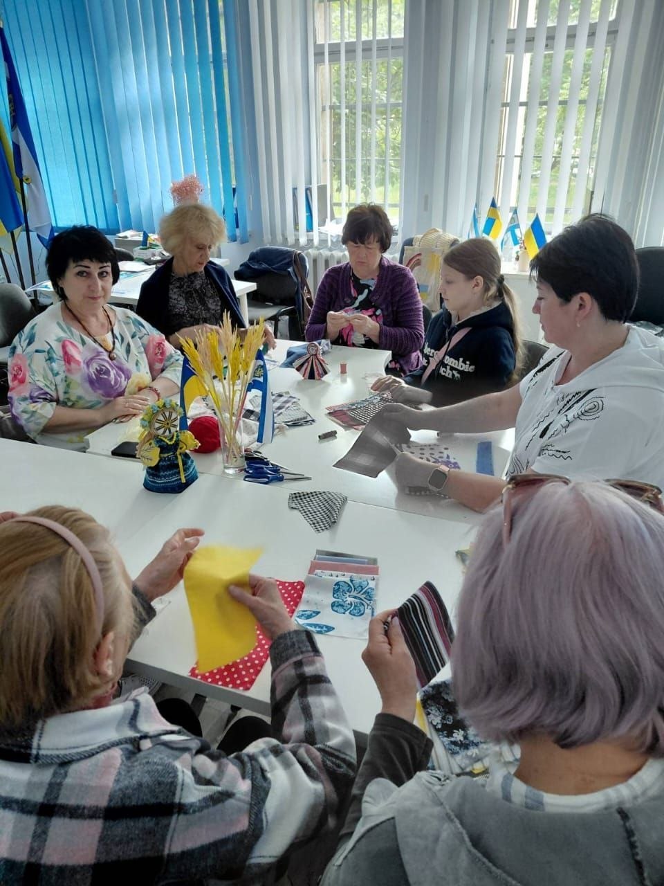 Група жінок різного віку та одна дівчинка сидять навколо білого столу у світлій кімнаті та займаються рукоділлям. Вони виготовляють традиційні ляльки-мотанки з тканинних клаптиків. На столі розкладено різнокольорові шматочки текстилю, нитки, ножиці, а також стоїть склянка з декоративними колосками пшениці та маленькими прапорцями України. Учасниці зосереджено працюють руками, дехто дивиться в кадр. Кімната має великі вікна з білими вертикальними жалюзі, а ліворуч на задньому плані видно частину синьо-жовтого прапора.