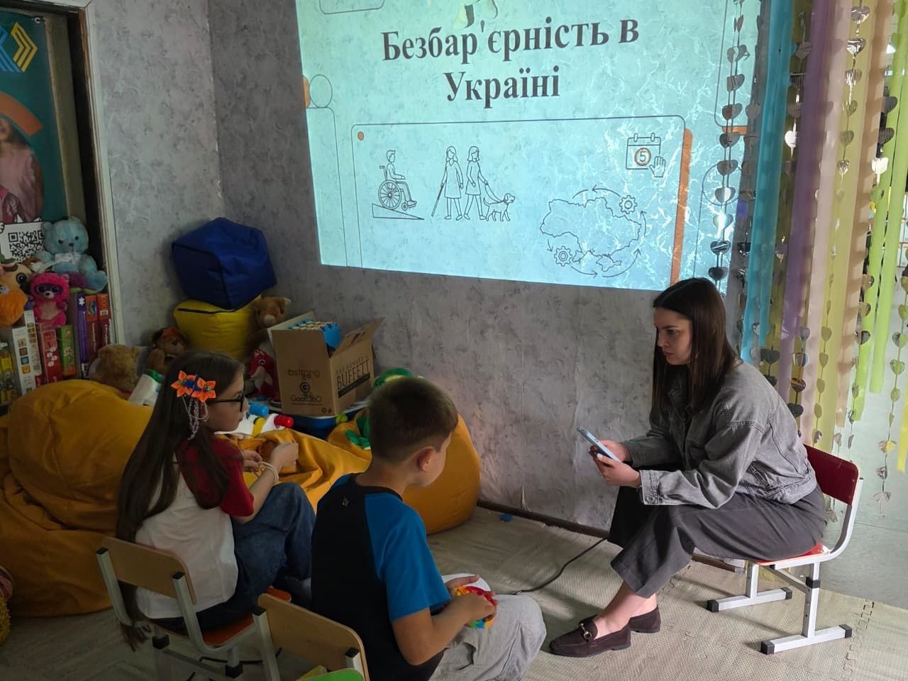 Заняття з дітьми про інклюзію. На стіну через проєктор виводиться слайд презентації з заголовком «Безбар'єрність в Україні» та лінійними малюнками людей на кріслі колісному та з собакою-поводирем. Перед екраном на пуфах сидять хлопчик і дівчинка, а навпроти них на низькому стільці сидіть лекторка з телефоном у руках, яка веде бесіду.
