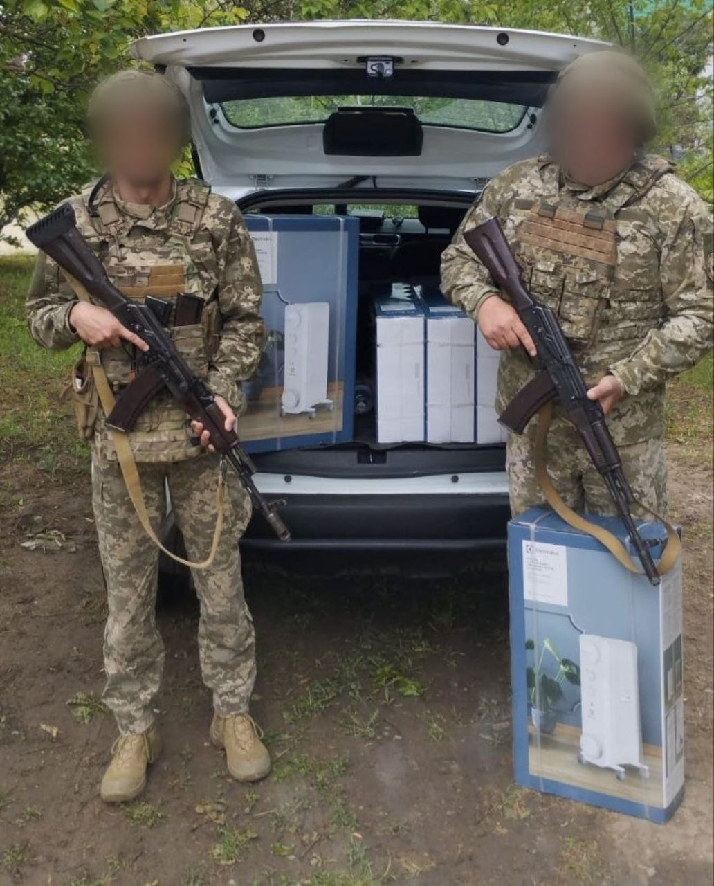 Двоє українських військових у камуфляжній формі, бронежилетах та касках стоять біля відчиненого багажника білого автомобіля. Їхні обличчя розмиті з міркувань безпеки. Обидва тримають у руках автомати. У багажнику машини лежать великі коробки з побутовою технікою, на яких зображені підлогові обігрівачі бренду Electrolux. Ще одна така коробка стоїть на землі праворуч.