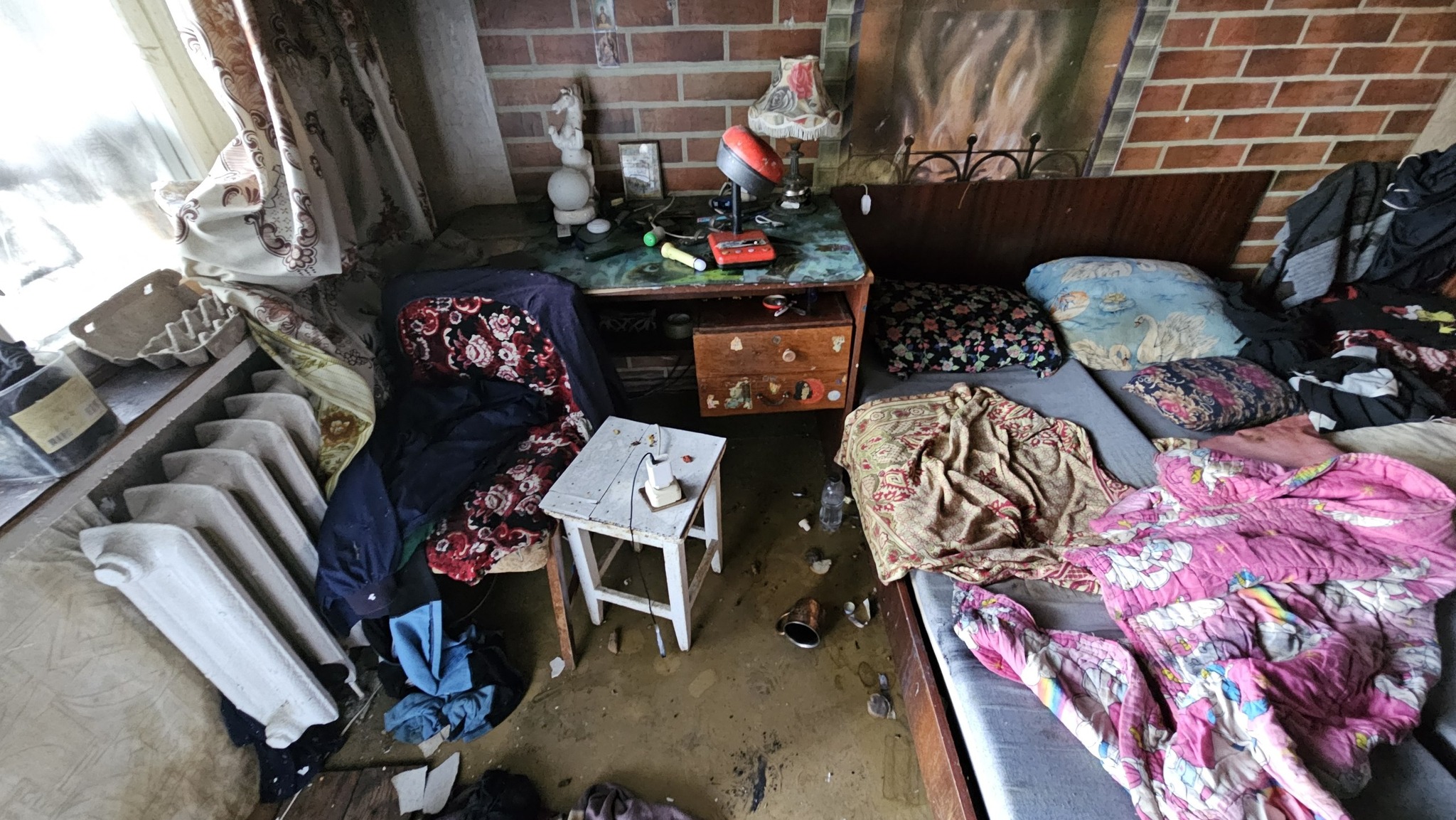 Житлова кімната, засипана уламками та пилом після вибухової хвилі або пожежі. На передньому плані ліворуч стоїть старий чавунний радіатор опалення, на стільці поруч розкиданий одяг. Справа розташоване дерев'яне ліжко з безладно пом'ятою рожевою ковдрою. На задньому плані видно письмовий стіл із настільними лампами та стіну, обкладену декоративною червоною цеглою. На підлозі помітні сліди води та бруду.
