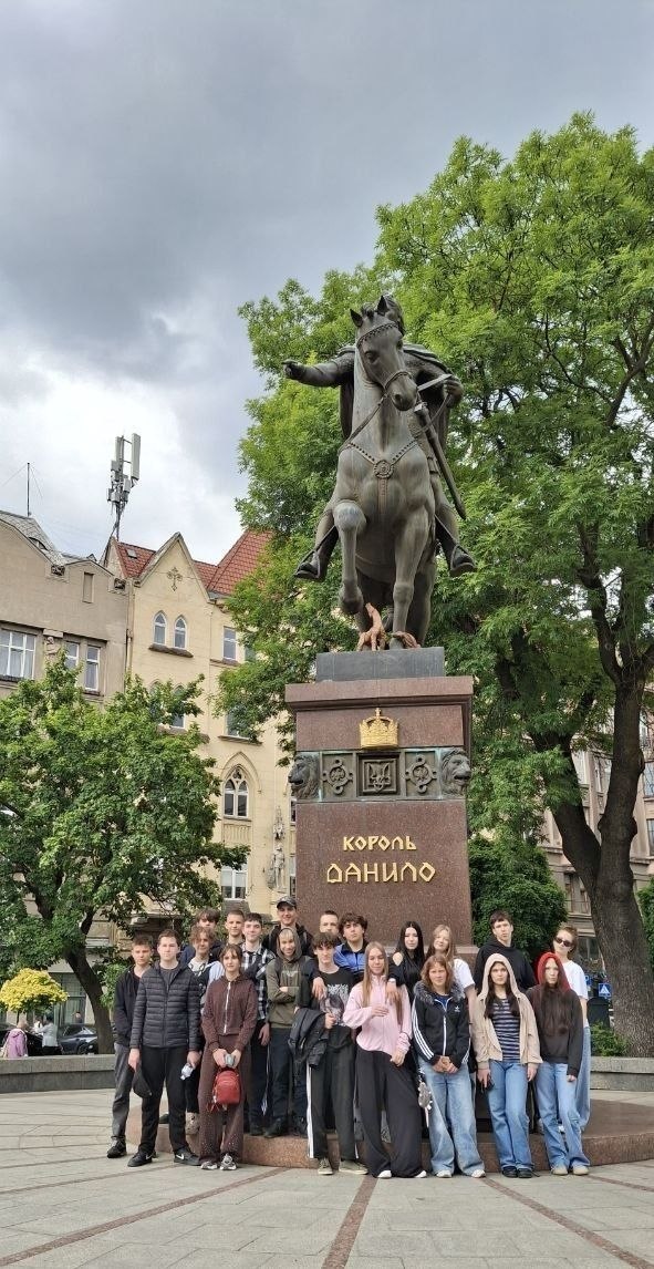 Альтернативний текст: Групова фотографія підлітків біля підніжжя пам'ятника Королю Данилу. Велика бронзова кінна статуя встановлена на високому кам'яному постаменті з написом «Король Данило». Близько п'ятнадцяти тинейджерів у повсякденному одязі стоять у ряд перед монументом на тлі зелених дерев та міської архітектури під хмарним небом.