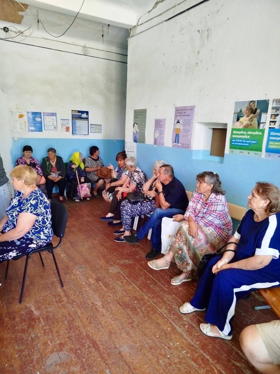 Черга з місцевих жителів, переважно літнього віку, у коридорі закладу охорони здоров'я чи адміністративної будівлі. Люди сидять на стільцях та лавках уздовж стін в очікуванні прийому. На світло-блакитних та білих стінах розвішані інформаційні плакати про вакцинацію та медичні послуги.