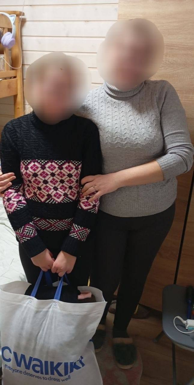 Жінка у сірому светрі під горло обіймає за плечі дівчинку в чорному в'язаному светрі з рожево-білим візерунком. Дівчинка тримає в руках великий білий фірмовий пакет із синім написом «Cwalkiki». Вони стоять у кімнаті біля дерев'яного двоярусного ліжка. Обличчя обох людей розмиті з міркувань приватності.