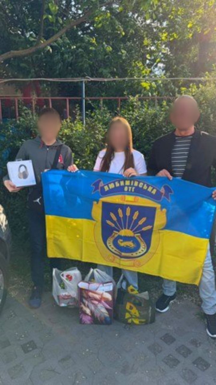 Двоє підлітків та чоловік тримають у руках великий синьо-жовтий прапор із написом «Любимівська ОТГ» та офіційним гербом громади (із зображенням підкови та колосків). Хлопець ліворуч тримає в руках коробку з навушниками. На землі біля ніг учасників стоять пакети та велика подарункова сумка з речами. Подія відбувається на вулиці біля кущів, обличчя людей приховані піксельним розмиттям.
