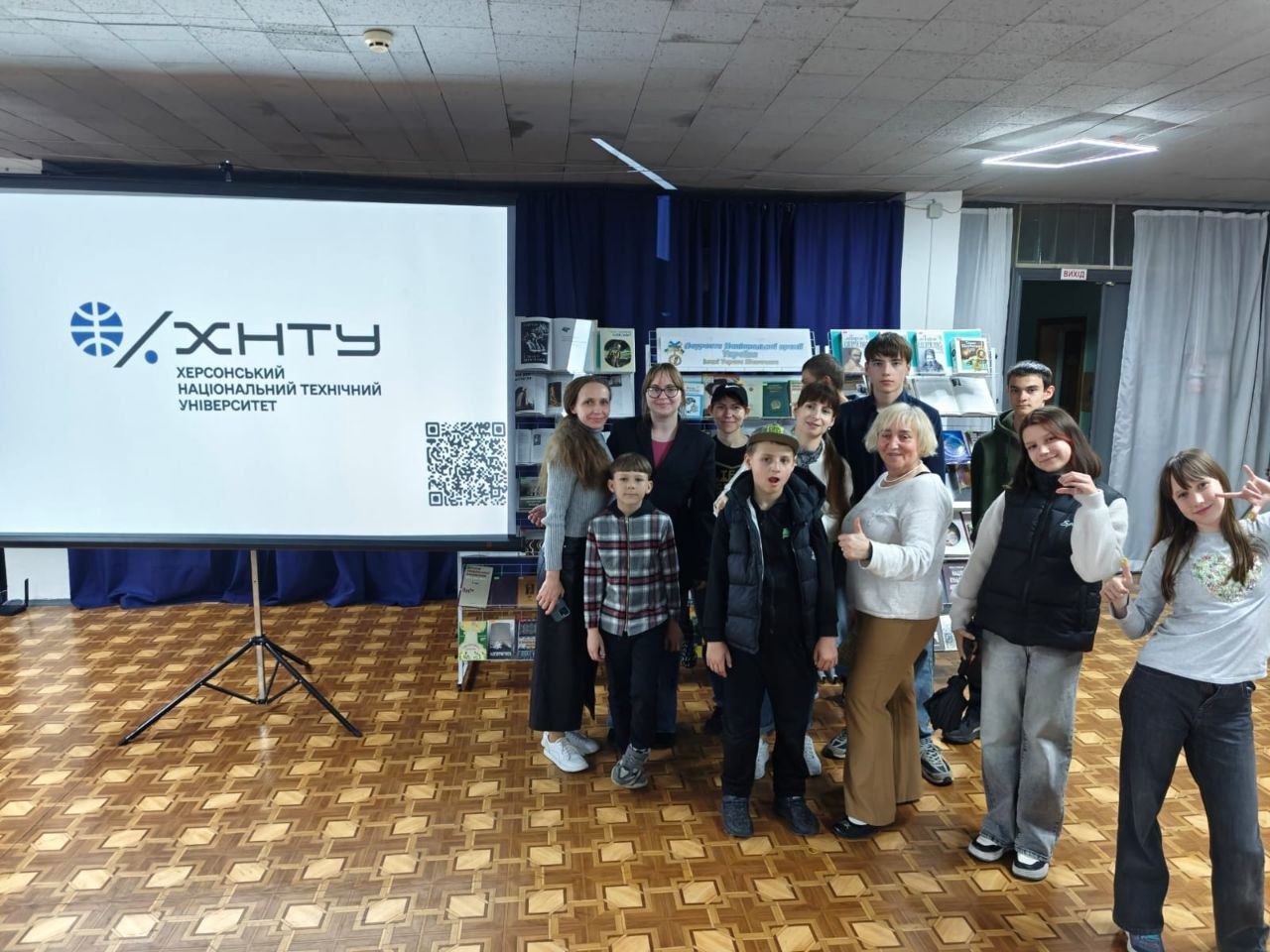 Групова фотографія викладачів та дітей у конференц-залі після заходу. Учасники стоять поруч із великим проєкційним екраном, на якому відображається логотип «ХНТУ — Херсонський національний технічний університет» та QR-код. На задньому плані видно книжкові стелажі з виставкою літератури.