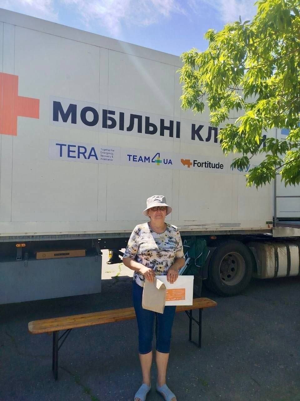 Жінка у світлій панамі, окулярах та квітчастій футболці стоїть перед великою білою вантажівкою мобільної клініки. В руках вона тримає паперовий пакет та інформаційні матеріали. На кузові машини нанесено великий червоний хрест, напис «МОБІЛЬНІ КЛІНІКИ» та логотипи партнерів, зокрема «TERA», «TEAM4UA» та «Fortitude». Поруч стоїть дерев'яна лавка.