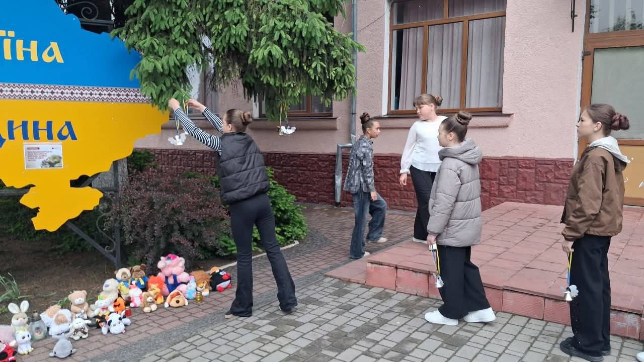Група дівчаток біля великого патріотичного стенда-карти «Україна ЄДИНА». Одна з учасниць акції у темній жилетці підняла руки вгору та прив'язує паперову фігурку білого ангела до нижніх гілок розлогої ялини, що росте поруч. Інші дівчатка з паперовими виробами в руках стоять поруч на сходах та тротуарі, очікуючи своєї черги. На землі під картою лежать м'які іграшки.