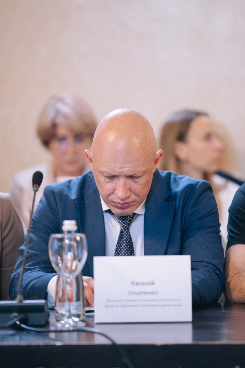 Портретний план заступника голови Херсонської обласної військової адміністрації Євгенія Ігнатенк. Чоловік із голеною головою, одягнений у синій піджак, білу сорочку та краватку, сидить за столом і зосереджено дивиться на документи перед собою. На передньому плані видно частину його іменної таблички та мікрофон. На розмитому задньому плані присутні інші учасники наради.