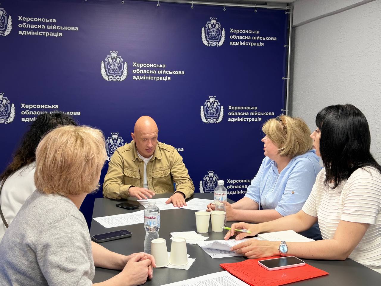 Альтернативний текст: Робоча зустріч у приміщенні Херсонської обласної військової адміністрації. За столом переговорів сидять п'ятеро людей — чоловік у тактичній куртці пісочного кольору та чотири жінки. Перед учасниками лежать документи, паперові стаканчики та пляшки з водою. На задньому плані — синій брендований банер із логотипами та написами «Херсонська обласна військова адміністрація».