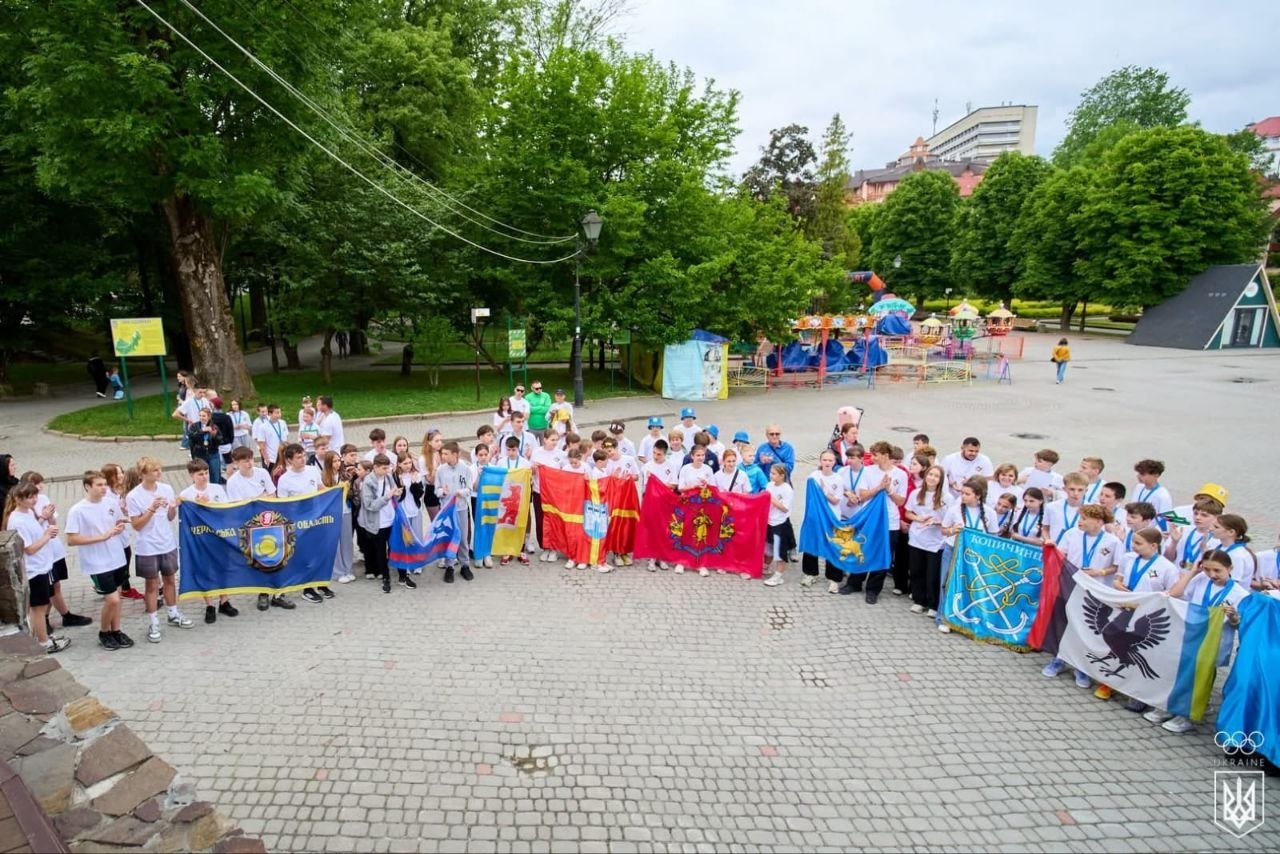 Загальний план з високої точки (ракурс згори). Велика група дітей-учасників, розбитих на команди, вишикувалася півколом на брущатій площі парку. Кожна команда тримає свій регіональний прапор або прапор громади (серед них видно прапори Черкаської області, Закарпаття, Копичинців тощо). Навколо — зелені дерева, елементи дитячого майданчика та міська забудова на задньому плані. Атмосфера офіційного закриття або відкриття всеукраїнських змагань.