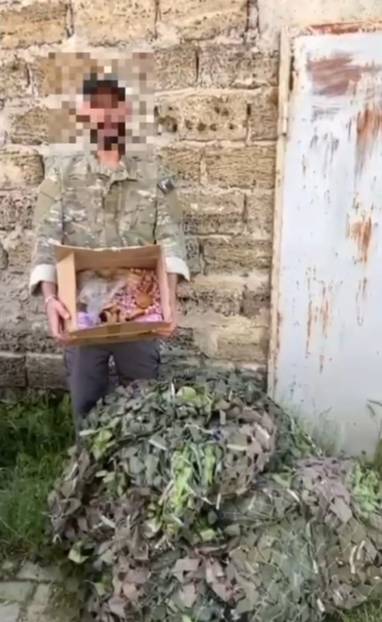 Чоловік у військовій формі тримає в руках картонну коробку з продуктами та іншими речами. Його обличчя приховане за допомогою піксельної цензури. Він стоїть на вулиці біля стіни з ракушняка та старих металевих дверей. Перед ним на землі лежить велика згорнута маскувальна сітка захисних зелених та коричневих відтінків.