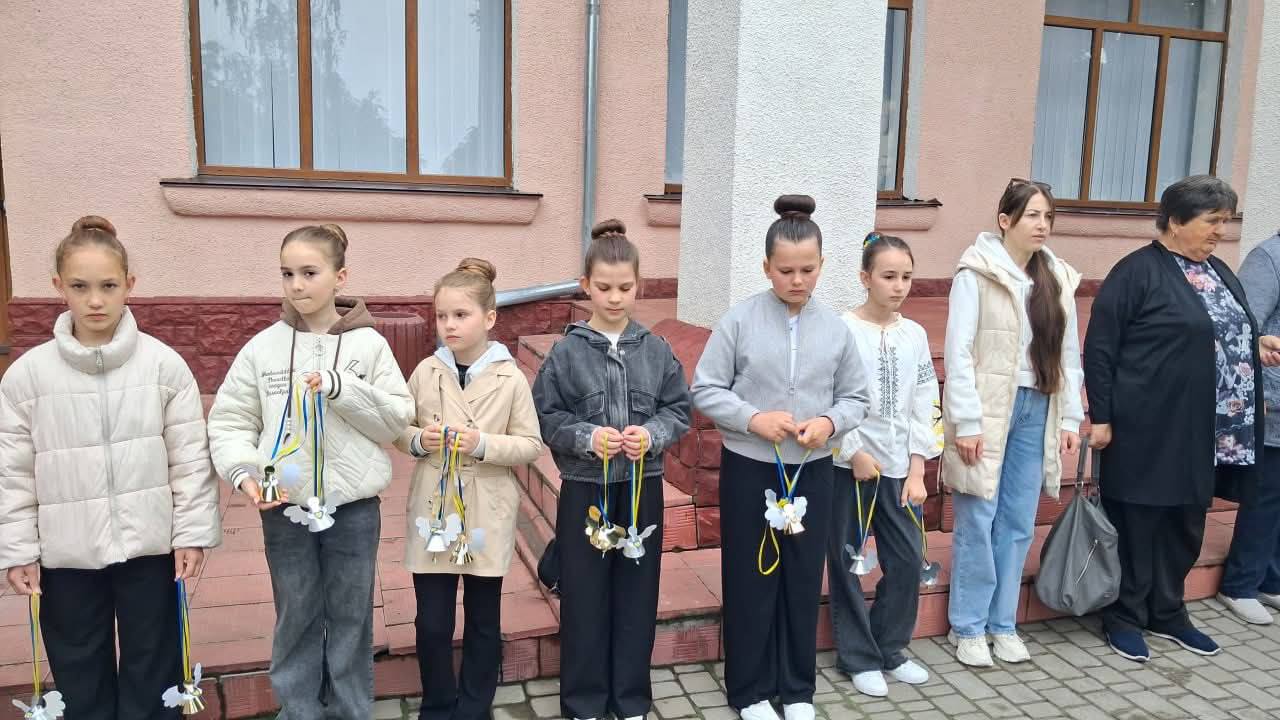 Шість дівчаток шкільного віку та дві дорослі жінки вишикувалися в ряд на вулиці вздовж стіни будівлі з великими вікнами під час офіційного заходу. П'ятеро дівчаток вдягнені у світлі куртки, кофти або вишиванку, тримаючи в руках виготовлені з паперу та металу дзвіночки на довгих синьо-жовтих стрічках. Усі присутні мають серйозний та зосереджений вираз обличчя.