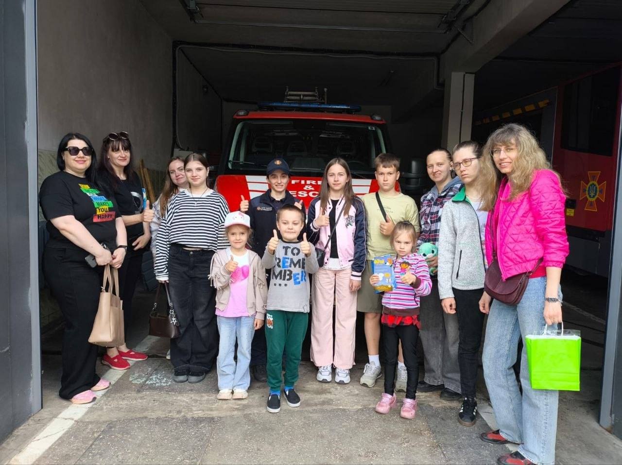 Групова фотографія дітей та дорослих у пожежному депо на тлі червоного рятувального автомобіля з написом «FIRE & ESCAPE». По центру стоїть співробітниця ДСНС у формі, а поруч із нею діти різного віку посміхаються та показують великий палець вгору. Праворуч видно частину іншої пожежної машини із символікою рятувальної служби.