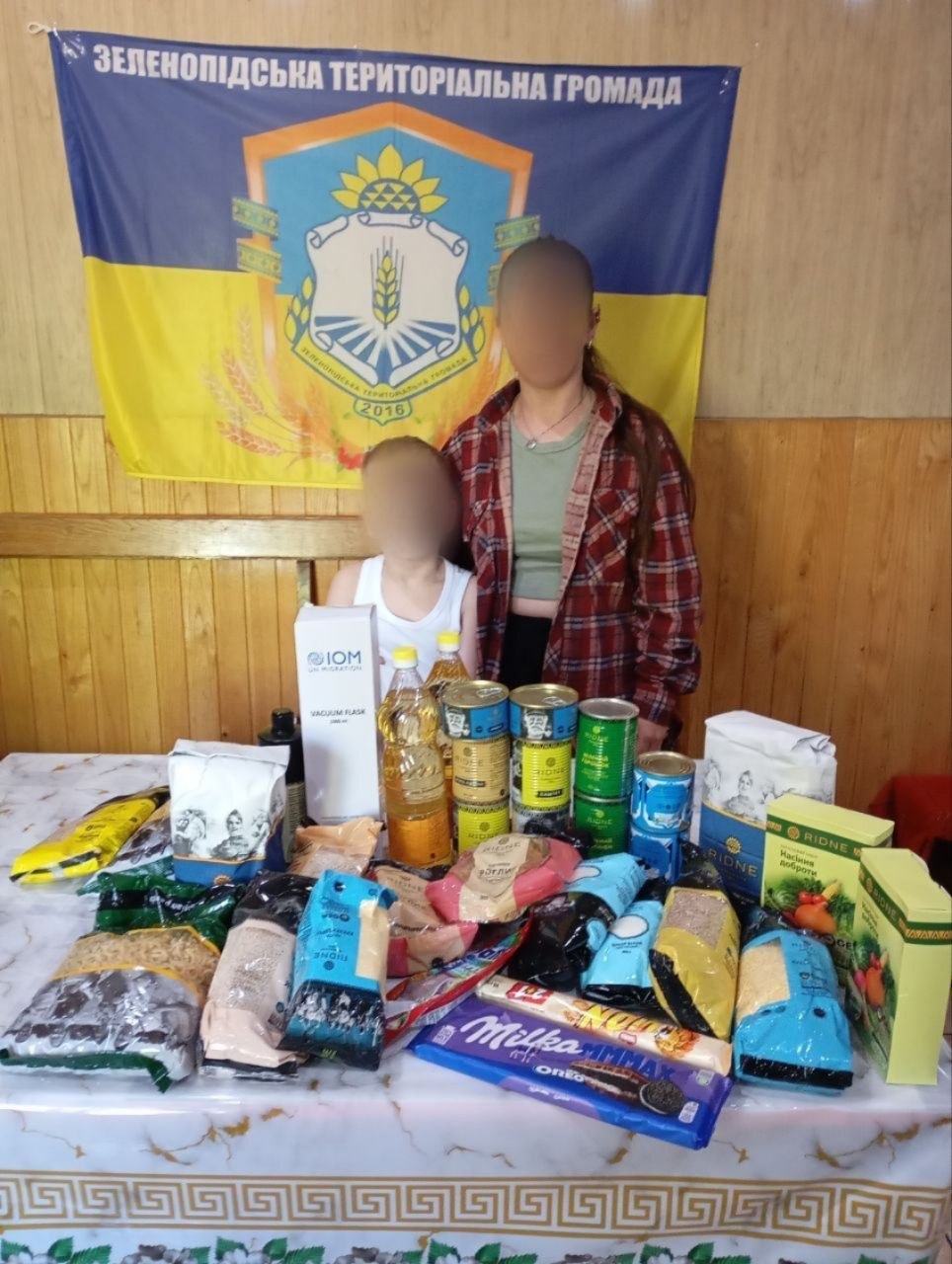 Жінка у картованій червоно-чорній сорочці та дитина у білій майці стоять за столом, на якому розкладено великий набір гуманітарної допомоги. Серед речей — коробка «IOM UN MIGRATION», пляшки з олією, бляшанки консервів, пакунки з крупами, макаронами, борошном, коробки з насінням «Насіння доброти» та велика плитка шоколаду Milka Max. На задньому плані на стіні висить синьо-жовтий прапор із написом «Зеленопідська територіальна громада» та гербом, датованим 2016 роком. Обличчя людей розмиті.