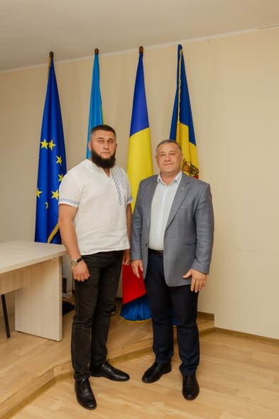 Офіційне фото двох чоловіків, які стоять поруч і тримають у руках сині папки з підписаними документами, на яких розміщено золотисті таблички з написами. Ліворуч — чоловік із бородою у білій вишиванці з синім орнаментом та чорних джинсах. Праворуч — чоловік у сірому піджаку та світлій сорочці. Вони стоять перед синім банером із жовтим написом «CONSILIUL ORĂȘENESC NISPORENI» та логотипом у вигляді млина. На столі праворуч видно маленькі прапори Європейського Союзу та Румунії.
