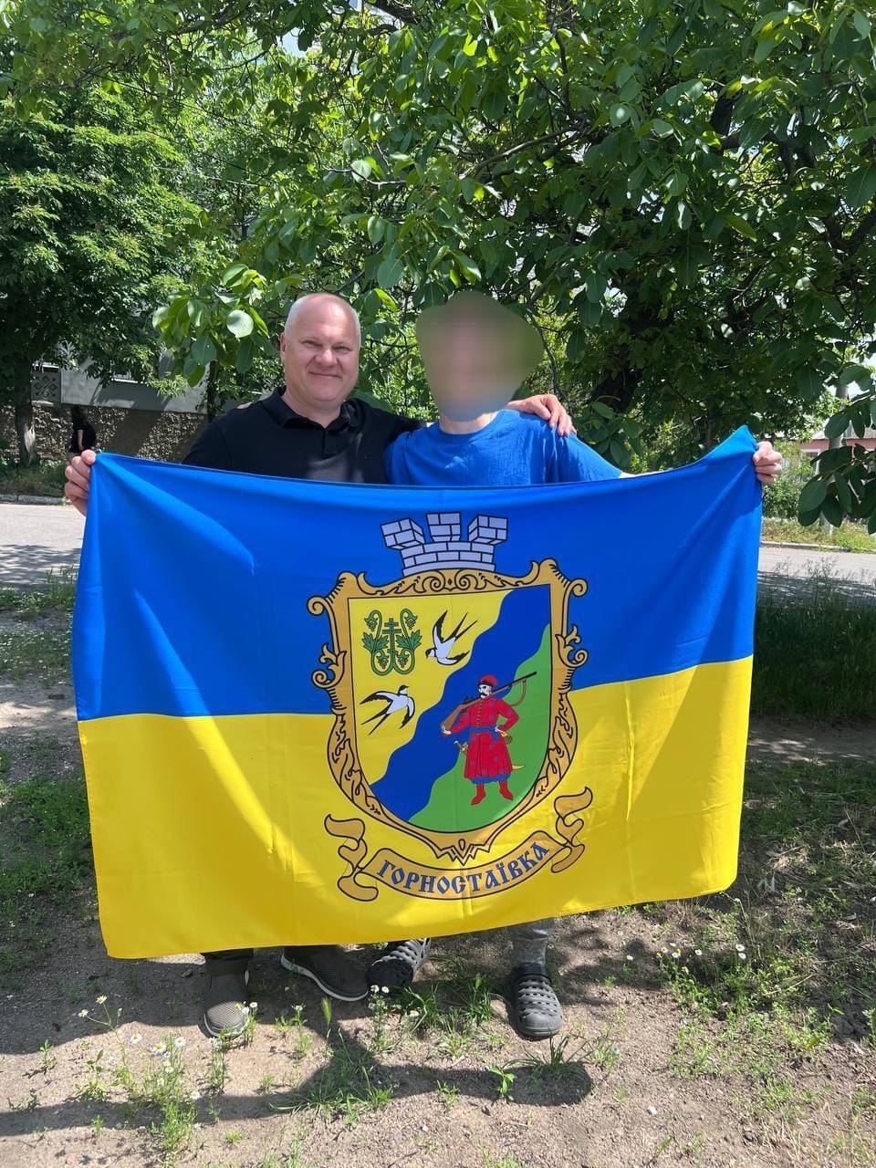 Чоловік у чорній футболці та підліток у синій футболці тримають великий синьо-жовтий прапор України з офіційним гербом селища Горностаївка. Вони стоять на ґрунтовій доріжці під розлогим зеленим деревом у сонячний день. Чоловік посміхається в камеру, обличчя підлітка повністю затушоване розмиттям.