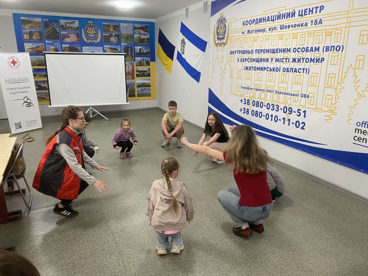 Психосоціальний тренінг та розваги для дітей-переселенців у житомирському координаційному центрі. П'ятеро дітей різного віку та дві волонтерки присіли навпочіпки по колу на підлозі й виконують спільну ігрову вправу. Ліворуч стоїть мобільний стенд «Психосоціальна підтримка» із символікою Червоного Хреста та білий проєкційний екран. На стіні праворуч розміщено великий інформаційний банер установи, а на дальній стіні — фотовиставка з краєвидами Херсонщини.