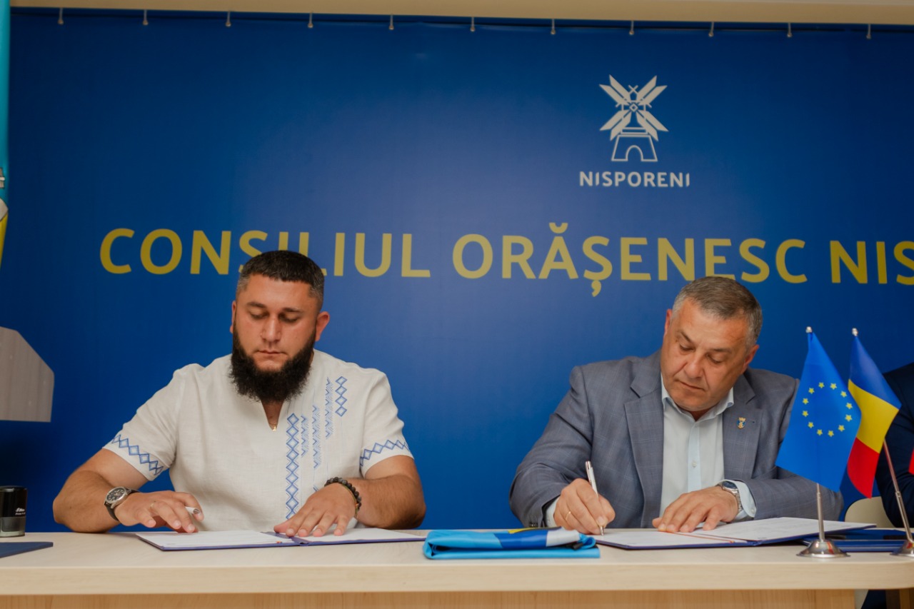 Процес підписання двосторонньої угоди. Двоє чоловіків сидять за світлим робочим столом і зосереджено підписують документи у відкритих папках. На столі перед ними стоять маленькі настільні прапорці Євросоюзу та Румунії, а також лежить складений синьо-жовтий прапор України. На задньому плані — синій офіційний банер міської ради Ніспорен.