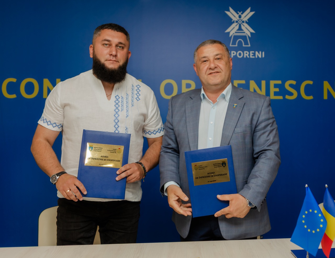 Офіційне фото двох чоловіків, які стоять поруч і тримають у руках сині папки з підписаними документами, на яких розміщено золотисті таблички з написами. Ліворуч — чоловік із бородою у білій вишиванці з синім орнаментом та чорних джинсах. Праворуч — чоловік у сірому піджаку та світлій сорочці. Вони стоять перед синім банером із жовтим написом «CONSILIUL ORĂȘENESC NISPORENI» та логотипом у вигляді млина. На столі праворуч видно маленькі прапори Європейського Союзу та Румунії.