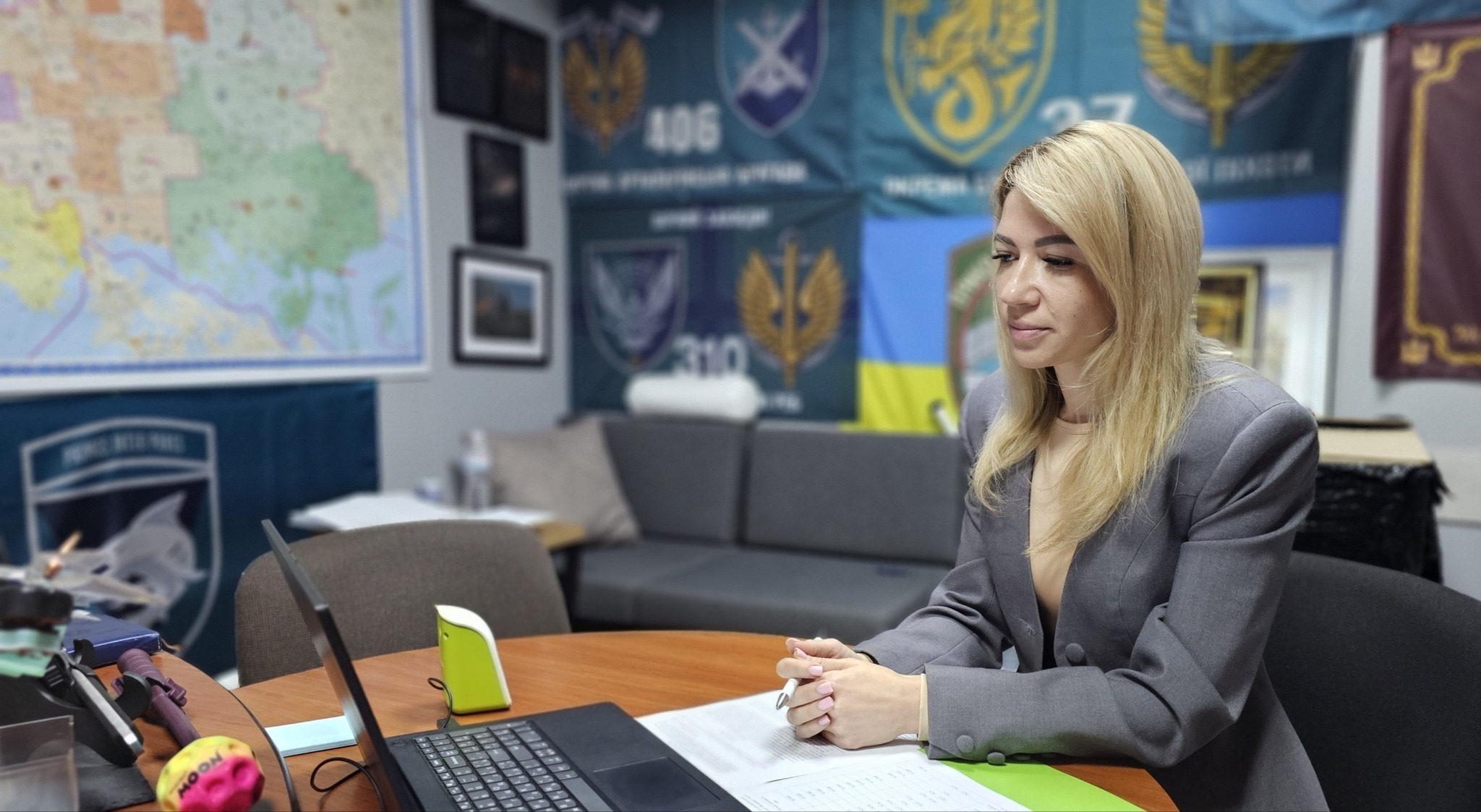 Жінка зі світлим волоссям у класичному сірому жакеті сидить за круглим дерев'яним столом у робочому кабінеті й бере участь в онлайн-нараді. Перед нею відкритий чорний ноутбук, робочі документи та ручка в руках. На задньому плані видно офісний диван, на стіні ліворуч висить велика географічна карта, а на стіні по центру — прапор України та численні військові прапори й банери підрозділів Сил оборони (зокрема з номерами 406, 310).