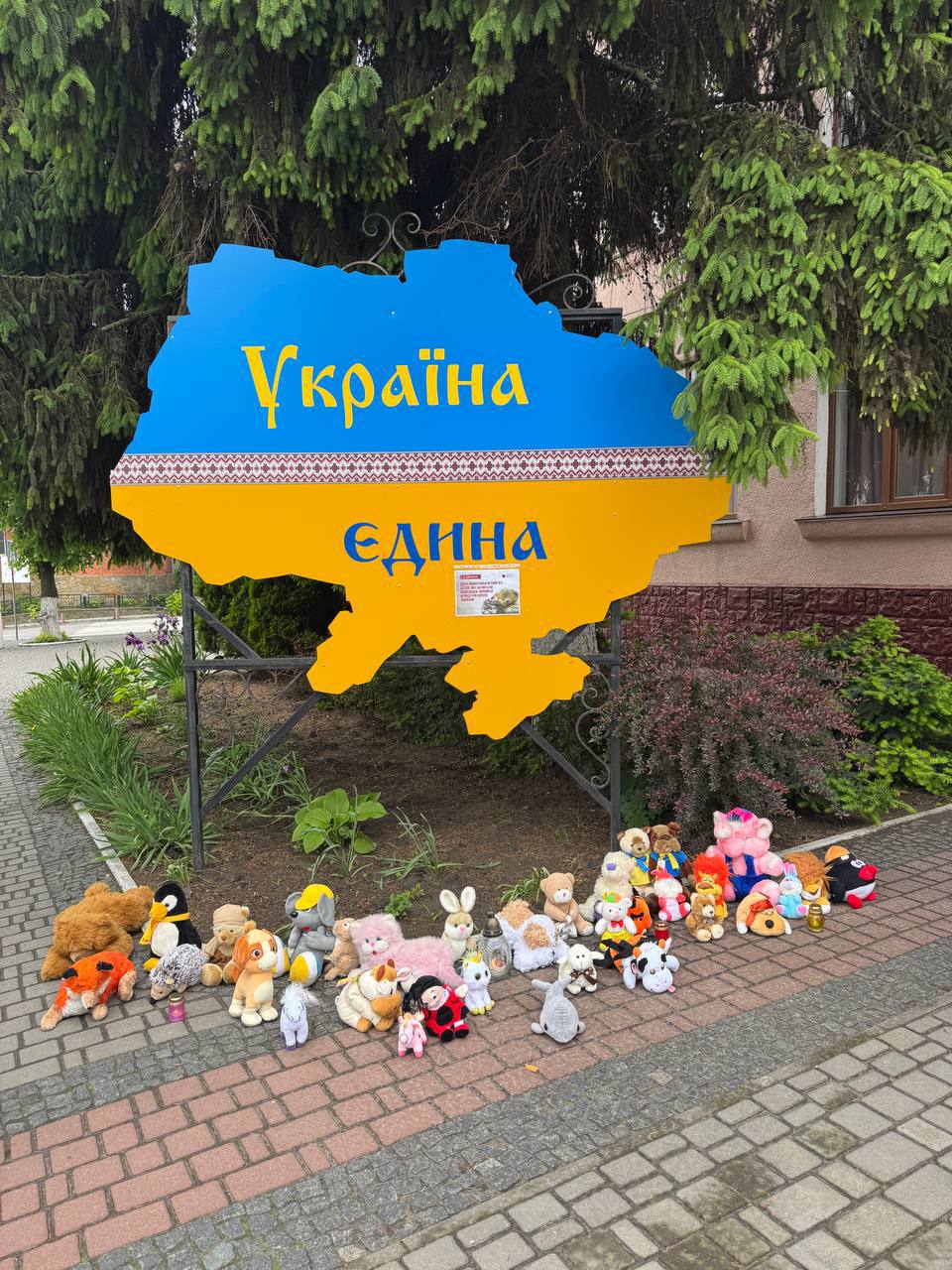 Великий вуличний стенд у формі карти України, пофарбований у кольори державного прапора — синій зверху та жовтий знизу. На синьому тлі нанесено жовтий напис «Україна», на жовтому — синій напис «ЄДИНА». Навколо металевої опори стенда, безпосередньо на тротуарній плитці біля газону, викладено в ряд кілька десятків дитячих м'яких іграшок (ведмедики, зайці, поросята), залишених як знак пам'яті. На задньому плані видно зелену хвою дерева та частину світлої офісної будівлі.