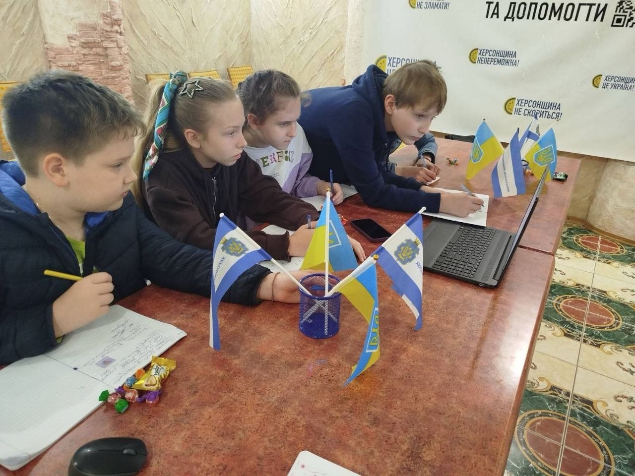 Група з чотирьох дітей сидить за довгим червоно-коричневим столом під час інтерактивного або освітнього заняття в хабі. Хлопчик на передньому плані ліворуч робить записи у зошиті, поруч із ним лежать кілька цукерок. Три інші дитини (дві дівчинки та хлопчик) зосереджено дивляться на екран відкритого чорного ноутбука, що стоїть праворуч. На столі у підставці та біля комп'ютера розставлені маленькі настільні прапорці України та міста Херсона. На стіні позаду видно написи «Херсонщина не скориться!» та «Херсонщина — це Україна!».