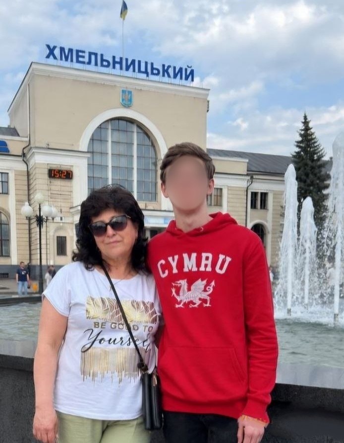 Жінка в сонцезахисних окулярах і білій футболці з написом «Be good to yourself» стоїть поруч із підлітком у яскраво-червоному худі з написом «CYMRU» та зображенням валлійського дракона. Вони позують на тлі будівлі залізничного вокзалу міста Хмельницький. Над входом до вокзалу встановлено великі сині літери «ХМЕЛЬНИЦЬКИЙ», а праворуч працює фонтан. На електронному табло вокзалу видно час 15:27. Обличчя хлопця розмите.