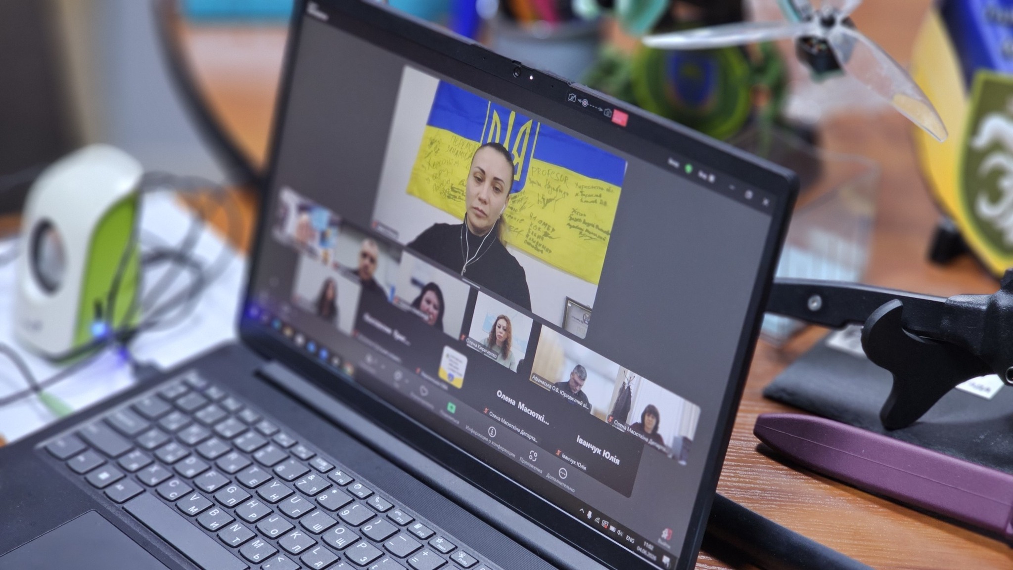 Крупний план екрана ноутбука, на якому запущено вікно відеоконференції Zoom або Microsoft Teams. Головне зображення на екрані показує жінку на тлі жовто-блакитного прапора України з підписами. Нижче в ряд розміщені вікна інших учасників наради, серед підписів видно імена «Олена Масюткі…» та «Іванчук Юлія». Ноутбук стоїть на робочому столі, поруч із ним розташовані комп'ютерна миша, дрібні деталі техніки та елементи безпілотника (квадрокоптера).