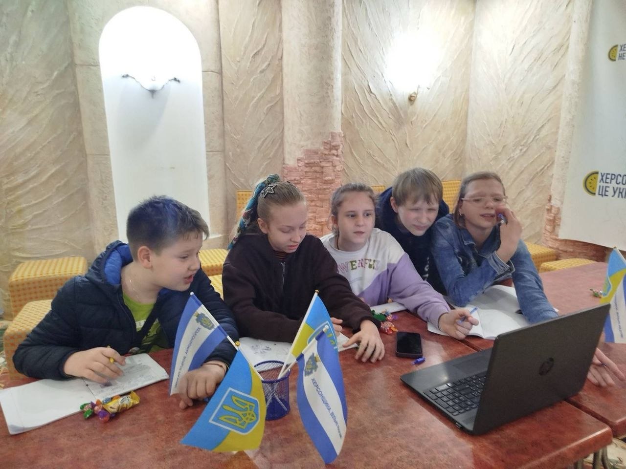 Вид з-за плечей дітей на екран ноутбука, за яким вони виконують логічне завдання або розгадують ребус. На моніторі відкрита таблиця з українськими літерами (А, Б, В, Г, Д тощо) та комбінаціями символів у лівій частині. На столі перед ноутбуком лежить чорний смартфон, зошити та кілька обгорток від цукерок. На задньому плані видно частину кімнати з розгорнутим державним прапором України та меморіальним або інформаційним банером для внутрішньо переміщених осіб (ВПО) у місті Одеса за адресою вулиця Балківська, 35/1.