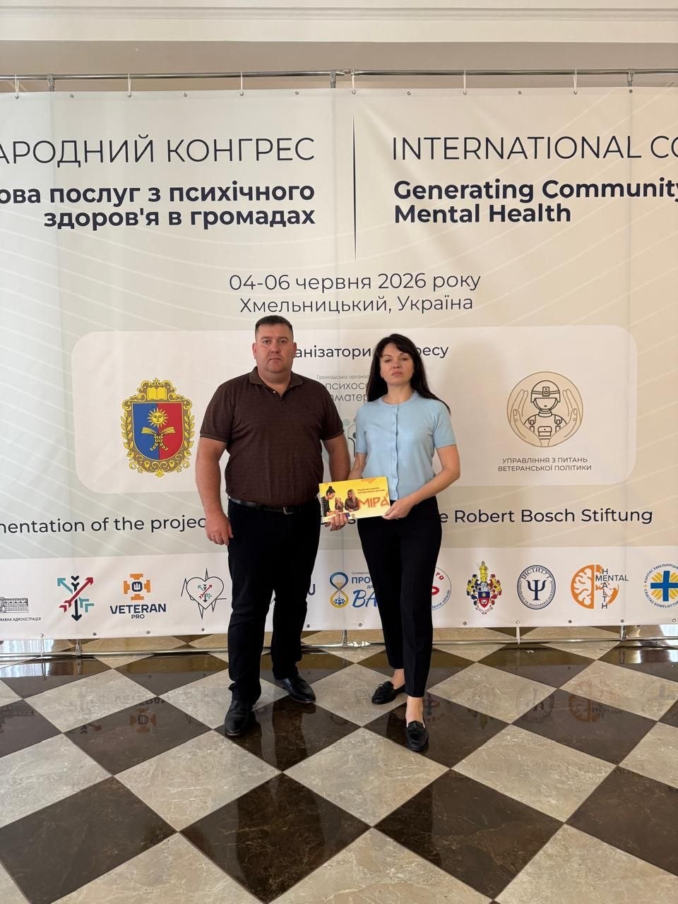 Чоловік у коричневому поло та жінка у блакитній блузці й чорних штанах позують у повний зріст на глянцевій підлозі з шаховим візерунком. У руках вони спільно тримають жовту інформаційну брошуру. Вони стоять перед великим офіційним банером заходу, на якому написано: «МІЖНАРОДНИЙ КОНГРЕС. Розбудова послуг з психічного здоров'я в громадах / INTERNATIONAL CONGRESS. Generating Community Mental Health. 04-06 червня 2026 року, Хмельницький, Україна». На банері також розміщено герб Хмельницької області, емблему Управління з питань ветеранської політики та логотипи партнерів, серед яких «Robert Bosch Stiftung» та «ВЕТЕРАН ПРО».