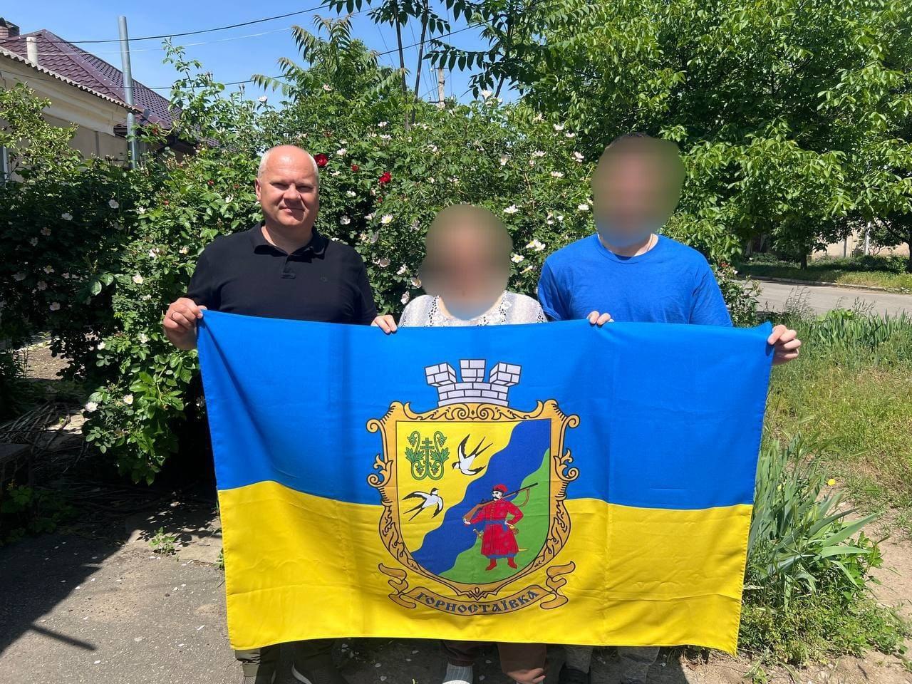 Двоє чоловіків та жінка стоять у повний зріст на вулиці на тлі зелених квітучих кущів рози й тримають розгорнутий державний прапор України, у центрі якого нанесено великий герб із написом «Горностаївка» (із зображенням козака з рушницею). Обличчя жінки та чоловіка праворуч у синій футболці розмиті, чоловік ліворуч у чорній футболці посміхається.