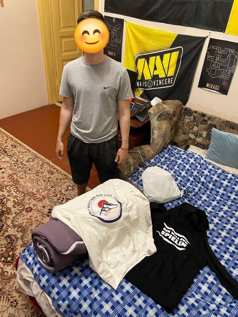 Молодий чоловік у сірій футболці Nike та темних шортах стоїть у кімнаті біля дивана, на якому розкладено нові речі. На синьо-білому картатому пледі лежать згорнута фіолетова ковдра, біле худі з круглим логотипом «Mountain Rafting» та чорне худі з написом «Der will nur spielen». На стіні позаду нього висить жовто-чорний прапор кіберспортивної команди «NAVI (Natus Vincere)» та ігрові постери з картами «Dust 2» і «Mirage».