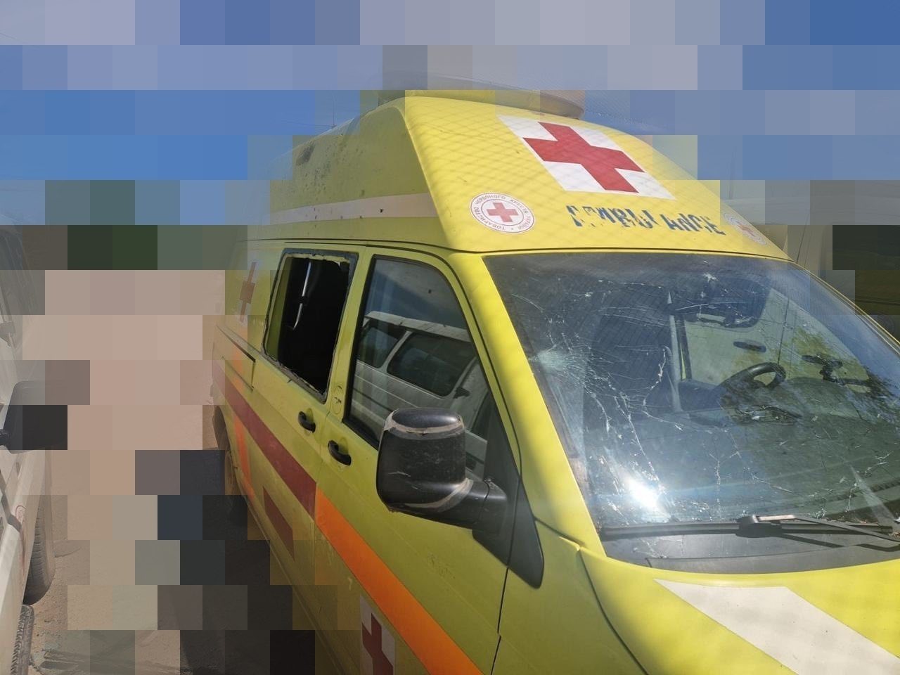 Жовтий автомобіль швидкої медичної допомоги (реанімобіль) з ознаками руйнувань. Лобове скло транспортного засобу сильно посічене тріщинами від уламків, а бокове скло водійських дверей повністю відсутнє (вибите). На високому даху машини нанесено великий червоний хрест та напис «AMBULANCE».