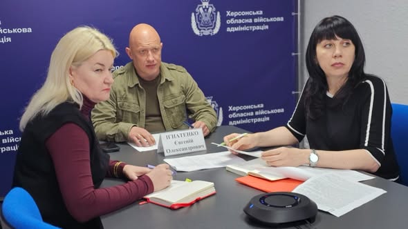 Робоча зустріч у приміщенні Херсонської обласної військової адміністрації на тлі синього брендованого банера ХОВА. За сірим столом сидять троє людей: ліворуч — жінка зі світлим волоссям у бордовому гольфі, яка робить записи в блокноті; по центру — лисий чоловік у тактичній куртці кольору хакі; праворуч — жінка з темним волоссям у чорній кофті, яка тримає ручку над документами. На столі лежать робочі папери, настільна табличка з ім'ям «Ігнатенко Євгеній Олександрович» та круглий чорний спікерфон для конференц-зв'язку.