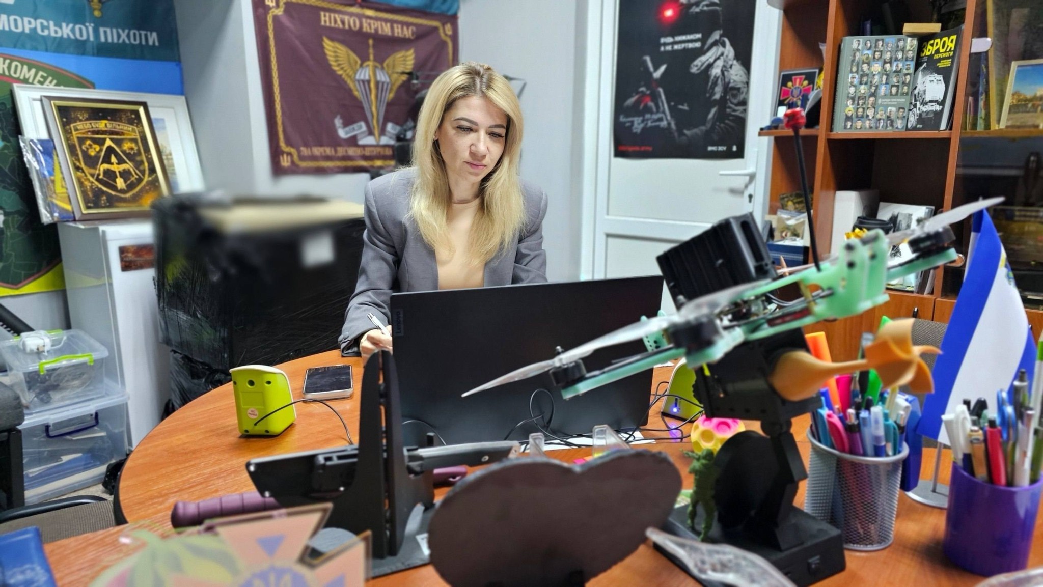 Жінка зі світлим волоссям у сірому блейзері сидить за круглим дерев'яним столом у робочому кабінеті й працює за ноутбуком. На передньому плані столу розташовані елементи військово-технічного спорядження: зібраний квадрокоптер зі світло-зеленими деталями, макети боєприпасів та підставки під канцелярію з ручками. На задньому плані, на стіні кабінету, висить бордовий прапор із військовою символікою та гаслом «НІХТО КРІМ НАС», мілітарі-плакати, а праворуч стоїть шафа з книгами та синьо-білим настільним прапорцем.