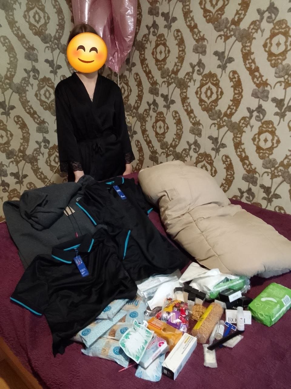 Дівчина у чорному халаті стоїть біля ліжка, на якому розкладено великий набір гуманітарної допомоги. Серед отриманих речей — велика бежева подушка або ковдра, дві чорні футболки поло з блакитною та синьою обробкою, сіра кофта, пакунки з гігієнічними засобами, упаковка печива, солодощі та медикаменти в коробках. Тло — стіна з класичними коричневими шпалерами з візерунком.
