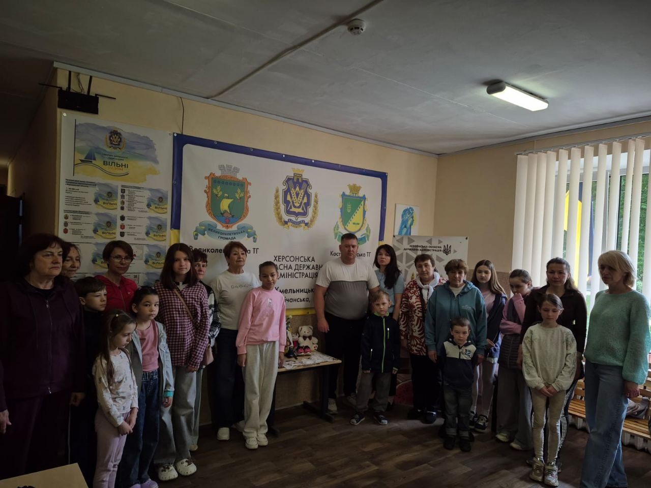Велика групова фотографія учасників заходу в приміщенні координаційного центру. Близько двадцяти людей — переважно жінки, підлітки та маленькі діти — вишикувалися для спільного знімка. Вони стоять на тлі великого білого банера з гербами Великолепетиської та Костянтинівської територіальних громад та написом «Херсонська обласна державна адміністрація». Ліворуч на стіні також розміщено інформаційні матеріали центру допомоги «Вільні разом».