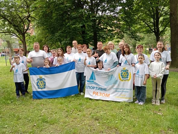 Велика групова фотографія учасників та організаторів сімейного спортивного заходу на зеленій галявині. Близько двадцяти людей вишикувалися в ряд. Зліва вони тримають великий синьо-біло-синій прапор міста Херсона з офіційним гербом. Справа учасники тримають білий банер із логотипом Херсонської ОВА, малюнком вітрильника та написами: «СПОРТИВНІ РАЗОМ» і «Гуманітарний Центр 