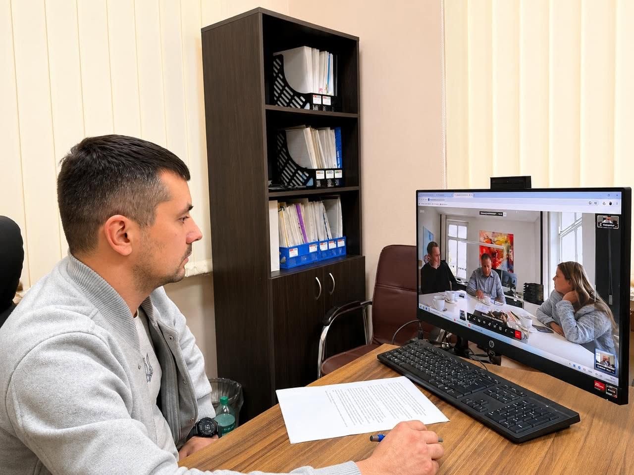 Чоловік у сірій кофті зі спортивним годинником на руці сидить за робочим столом у кабінеті в профіль до камери. Він бере участь в онлайн-нараді, дивлячись на великий монітор комп'ютера HP. На екрані відображається вікно відеоконференції, де видно трьох інших учасників (двох чоловіків та дівчину), які сидять за столом у світлій кімнаті. Перед монітором на дерев'яному столі лежать чорна клавіатура, аркуш паперу та ручка. На задньому плані стоїть темна офісна шафа з папками для документів.