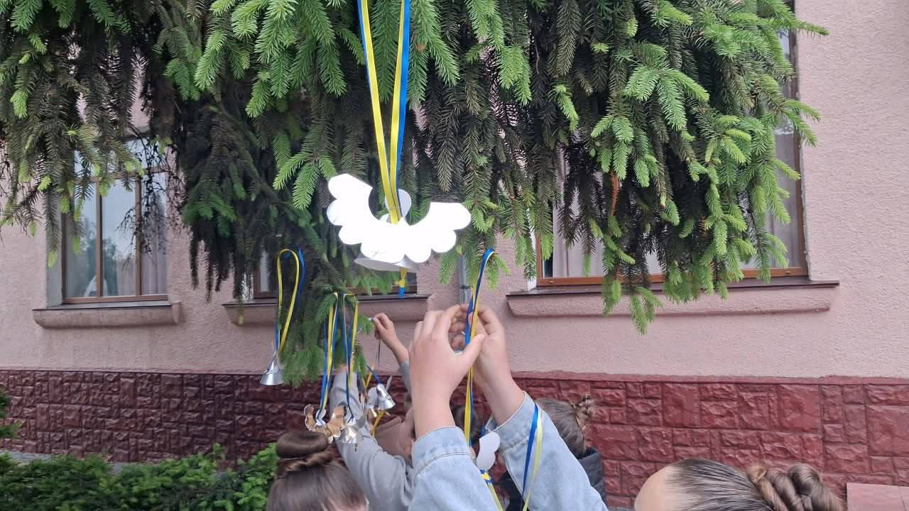 Крупний план дитячих рук, які піднімають вгору та закріплюють на нижніх зелених гілках хвойного дерева саморобні вироби — білих паперових ангеликів та блискучі металеві дзвоники. Кожен виріб підвішений на тонких атласних стрічках синього та жовтого кольорів. На задньому плані видно рожеву штукатурку стіни та коричневі декоративні панелі будівлі.