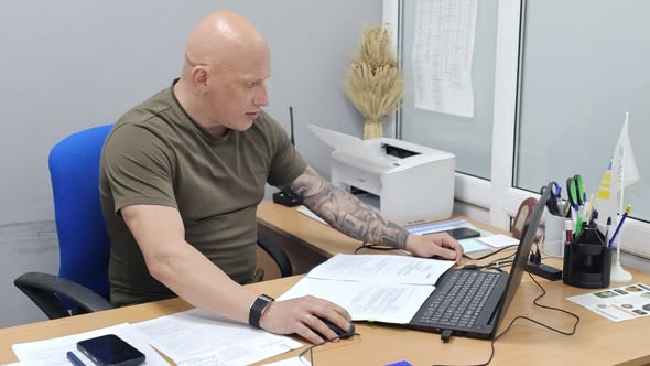 Середній план: лисий чоловік з татуюванням на правій руці, одягнений у футболку кольору хакі, сидить за офісним столом на синьому кріслі. Він кладе руку на роздруковані документи, дивлячись у бік. Перед ним відкритий ноутбук та папери. На столі видно принтер, канцелярське приладдя, невеликий прапорець і смартфон. Тло — світлі офісні стіни та вікно зі скляними панелями.